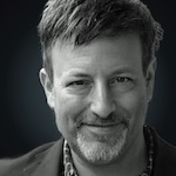 Black-and-white headshot of Chuck Brooks with short hair and a trimmed goatee, smiling gently. He is wearing a dark blazer over a checkered shirt, and the background is dark with a subtle gradient that highlights his face.