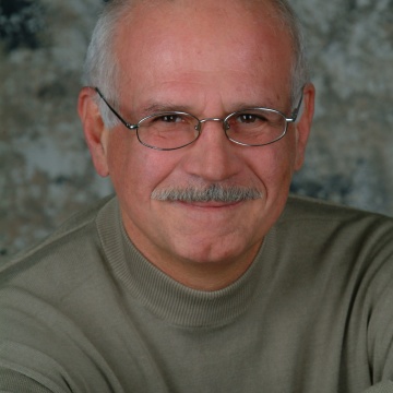 Headshot of Andrew Spanyi with short gray hair, a gray mustache, and wire-rimmed glasses, smiling gently at the camera. He is wearing a light olive-green sweater and is seated with his arms folded in front of him.