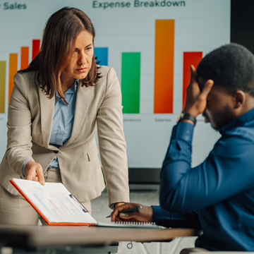 A manager reviews a report with a stressed colleague in front of a presentation displaying sales and expense charts.
