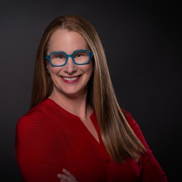 Professional headshot of Beth Rudden, CEO and co-founder of Bast AI, smiling with long, straight light brown hair wearing bright blue rectangular glasses and a red blouse. She is posed with her arms crossed against a dark gray studio background, facing the camera with a friendly, approachable expression.